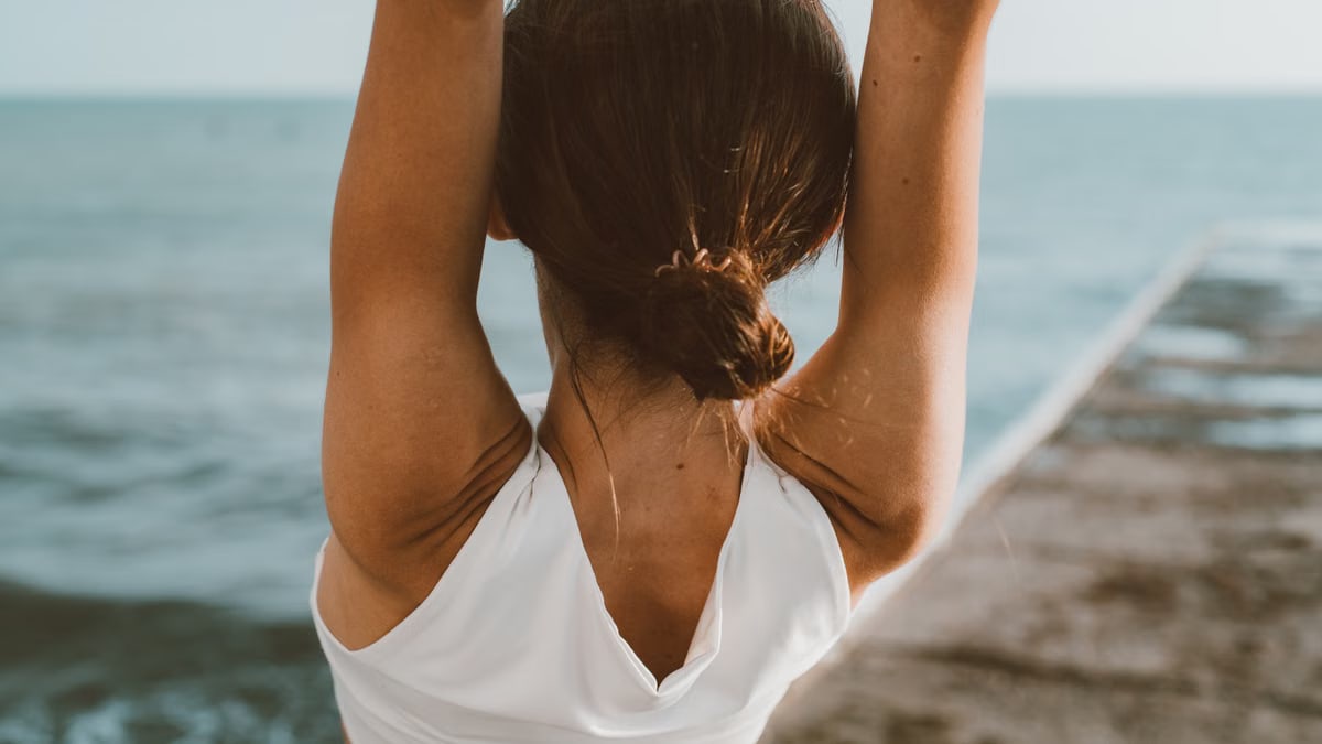 Back view of a woman meditating with raised hands by the ocean, symbolizing relaxation and a healthy lifestyle.