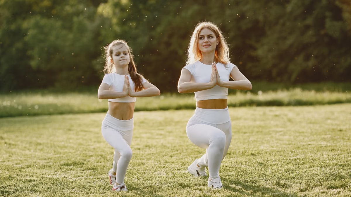 A mother and daughter practicing yoga together in matching outfits in a sunny park.